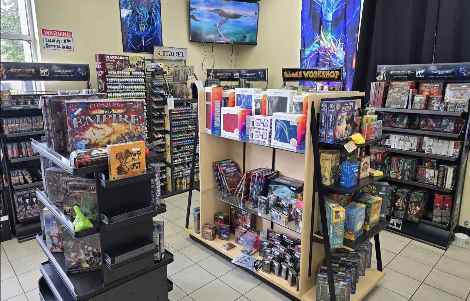 Inside The Dragon's Roost: shelves of Warhammer boxes, Citadel paints, board games, and card supplies, with open floor space between the displays.